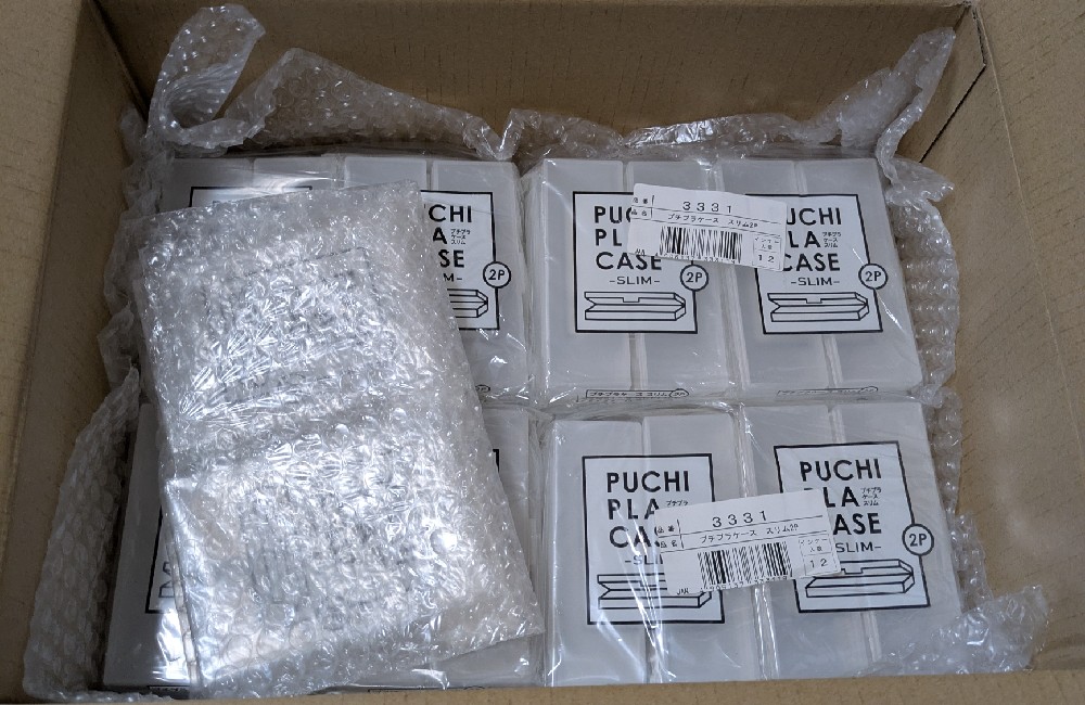 段ボール箱にきれいに並べられた、書類整理用の透明なプラスチック製の小物ケース。緩衝材で丁寧に梱包されており、行政書士事務所の几帳面な備品管理の様子。